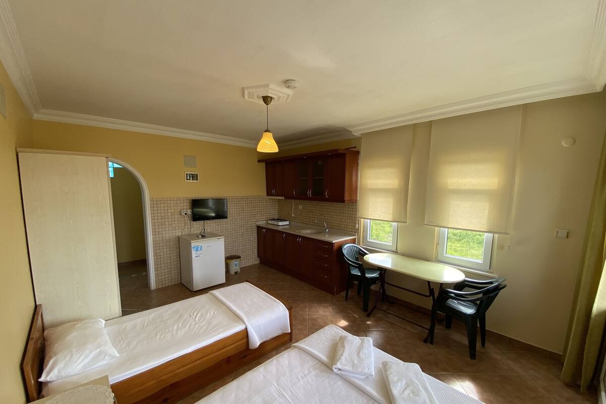 A compact room with two beds, a small kitchenette, a dining table with chairs, and a window with blinds.