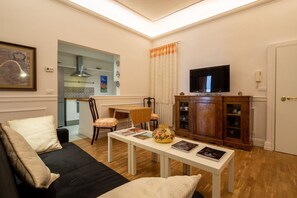 Una sala de estar con un sofá, una mesa con libros, un televisor sobre un mueble y una cocina al fondo.