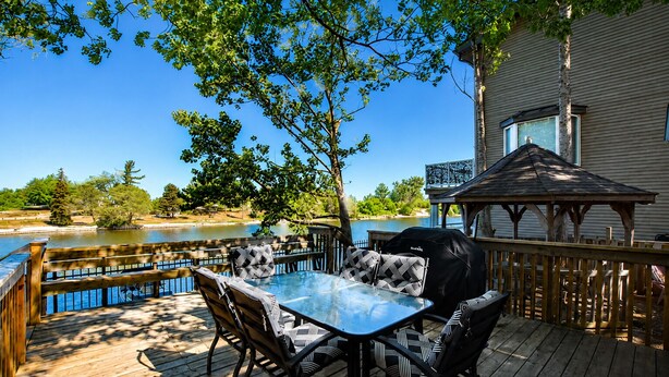 A deck with a glass-top table and chairs, a grill, and a view of a lake.
