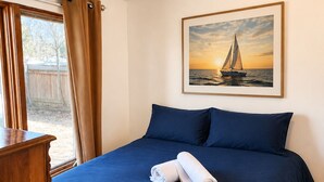A neatly made bed with a blue bedspread and pillows, a framed picture of a sailboat on the wall, and a wooden nightstand.