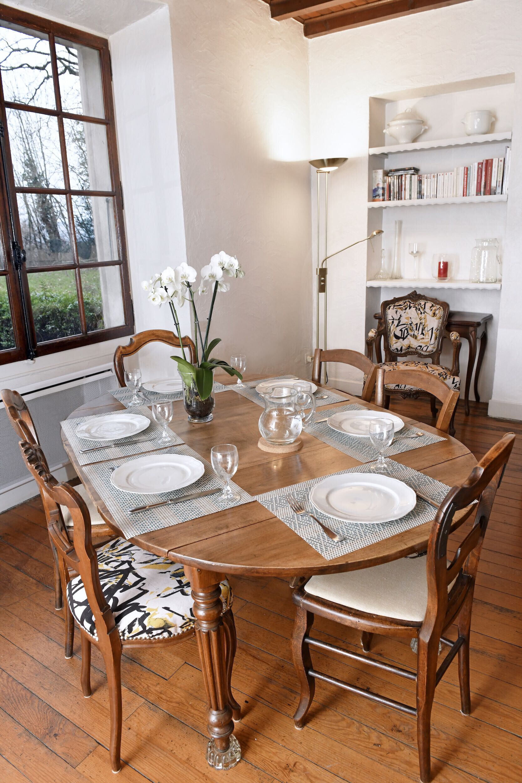 A dining room with a wooden table set for six, surrounded by six chairs with cushioned seats. A vase with white flowers is on the table, and a bookshelf with books and decorative items is in the background.