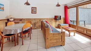 Ein gemütlicher Raum mit Holztisch, Bänken, einem Fernseher und einem großen Fenster mit roten Vorhängen.