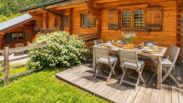 A wooden cabin with a dining table set outside, surrounded by greenery and a mountain backdrop.