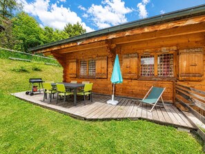 A wooden cabin with a deck, a table with chairs, a lounge chair, and a cart.