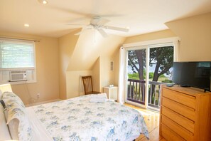 A bedroom with a bed, a dresser, a chair, a ceiling fan, a window with blinds, and a view of a tree and water outside.