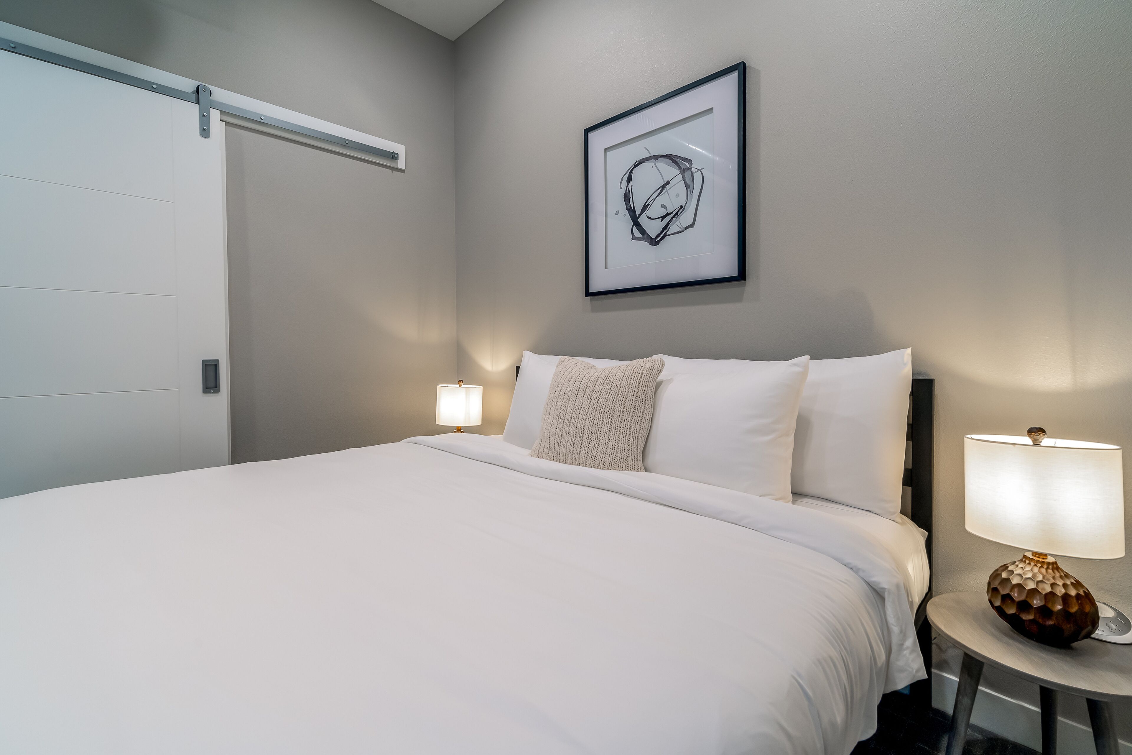 A neatly made bed with white linens, a framed abstract artwork above it, and two bedside lamps on either side.