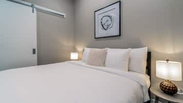 A neatly made bed with white linens, a framed abstract artwork above it, and two bedside lamps on either side.