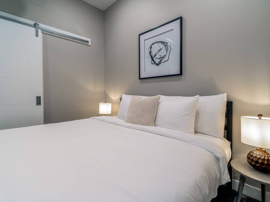 A neatly made bed with white linens, a framed abstract artwork above it, and two bedside lamps on either side.
