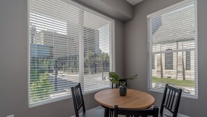 Een eetruimte met een ronde tafel en vier stoelen, grote ramen met zonwering en een potplant op de tafel.
