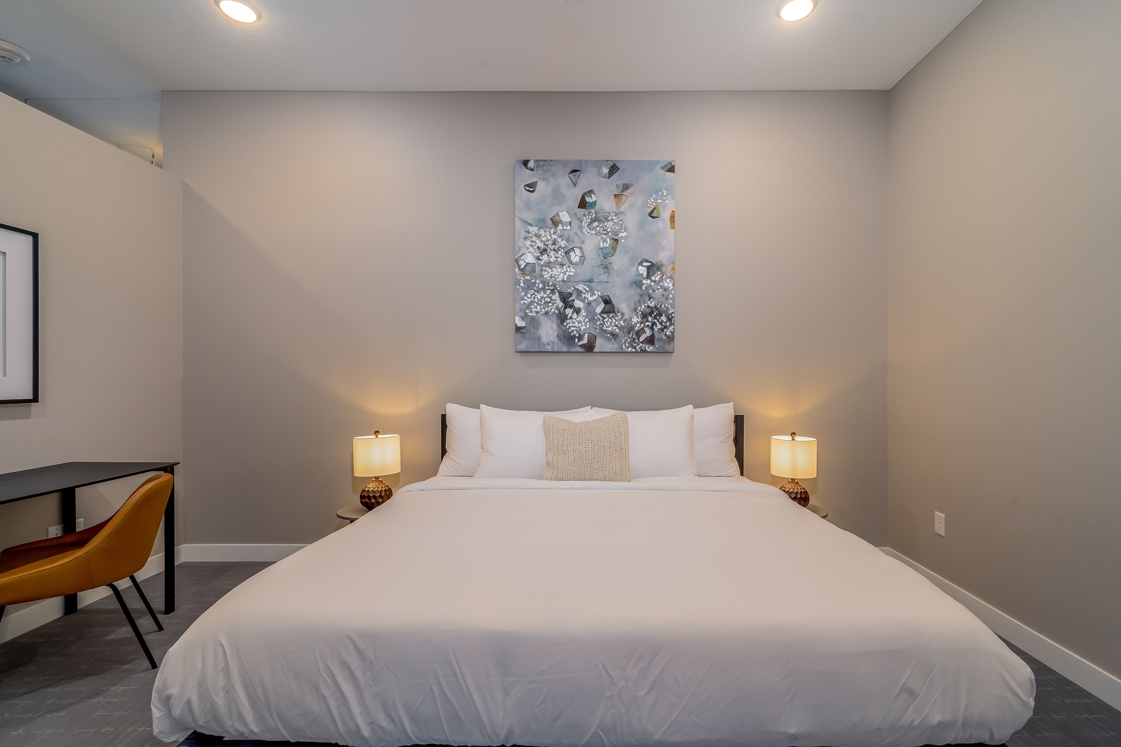 Habitación de hotel con una cama grande, dos lámparas de noche, un escritorio con silla y un cuadro abstracto en la pared.