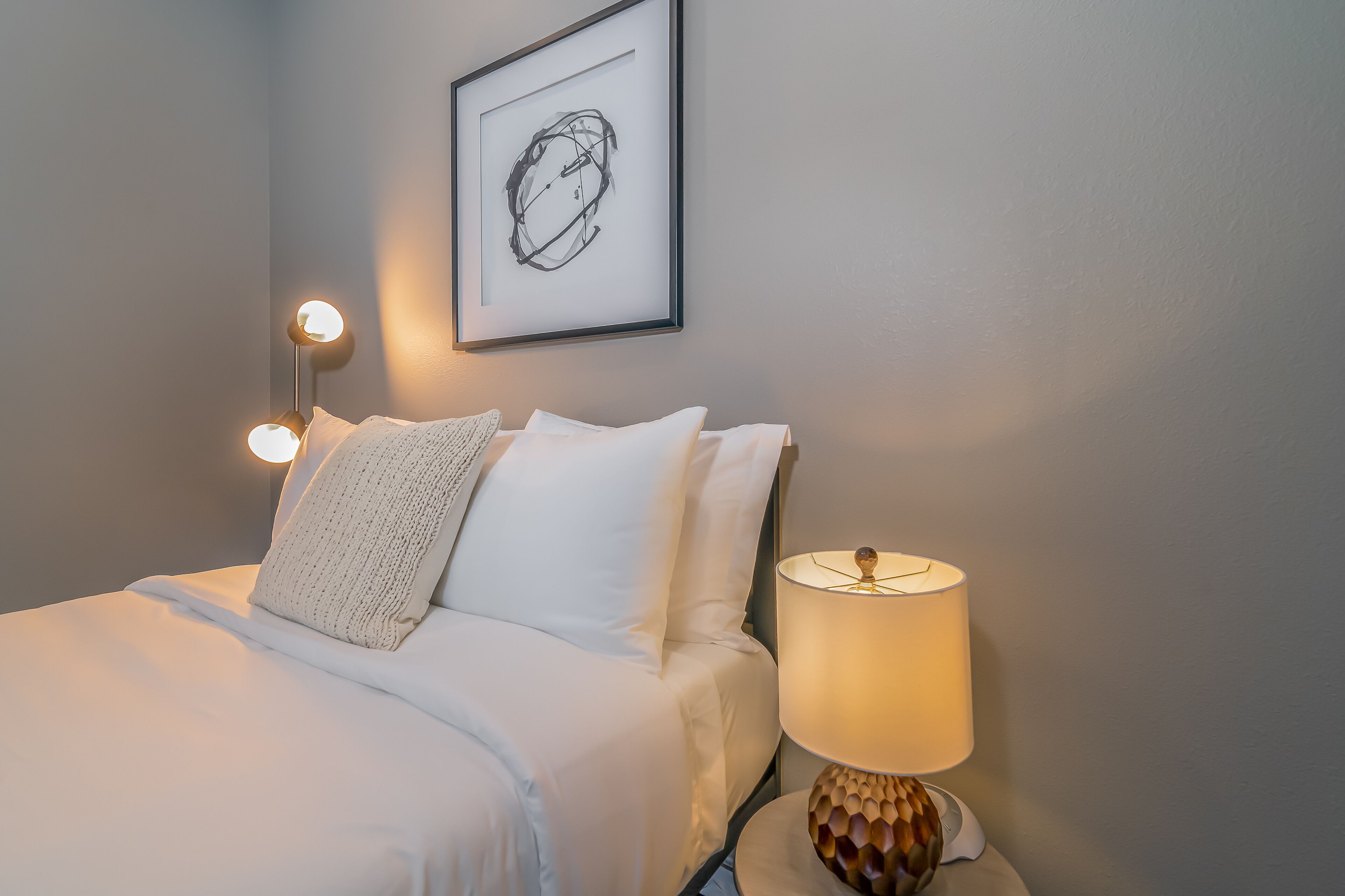 Un lit bien fait, avec du linge de lit blanc et un seul coussin décoratif, une œuvre d’art abstraite encadrée accrochée au mur, et une table de chevet avec une lampe et un téléphone.
