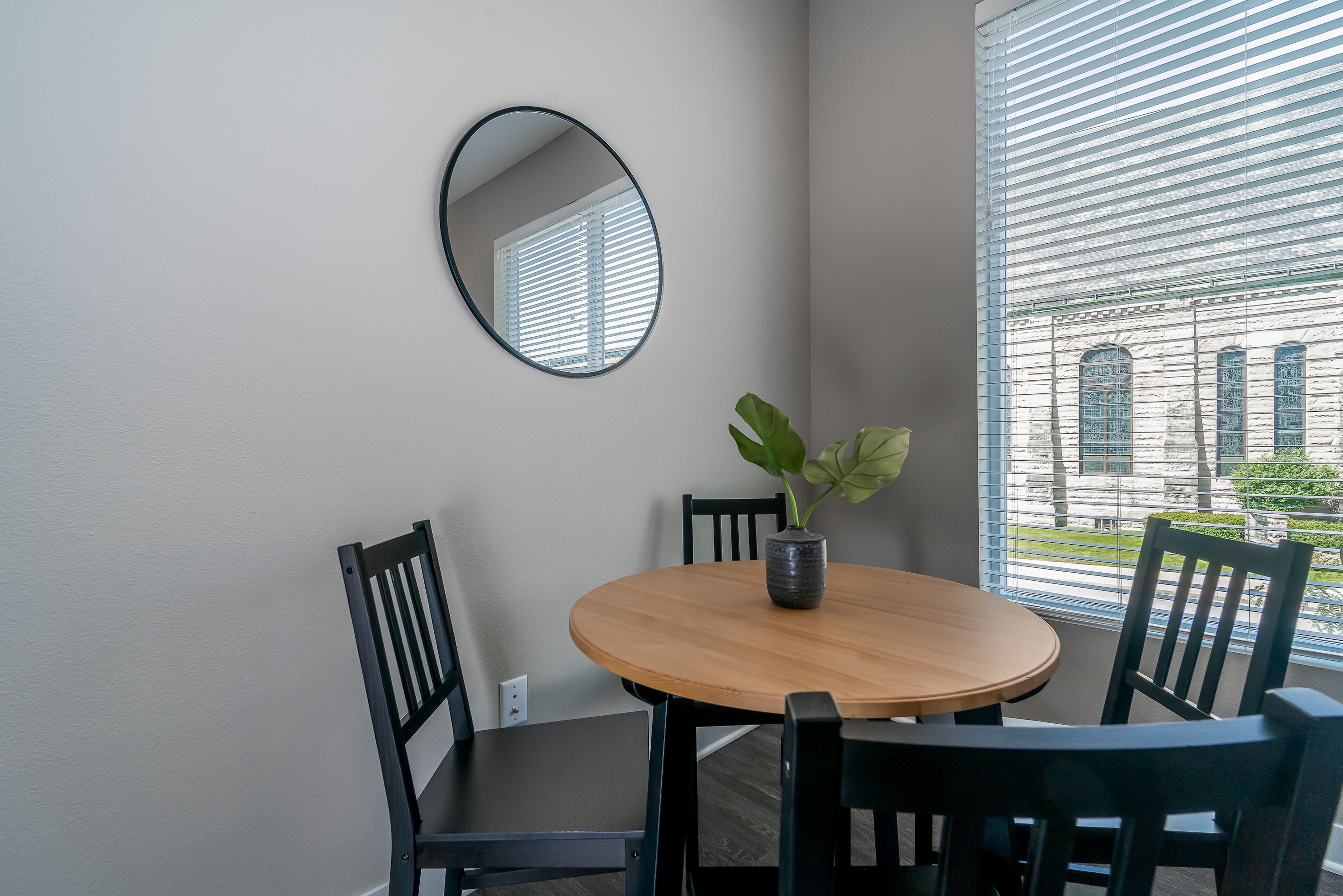 A dining area with a round wooden table, two black chairs, a potted plant, and a wall-mounted mirror.