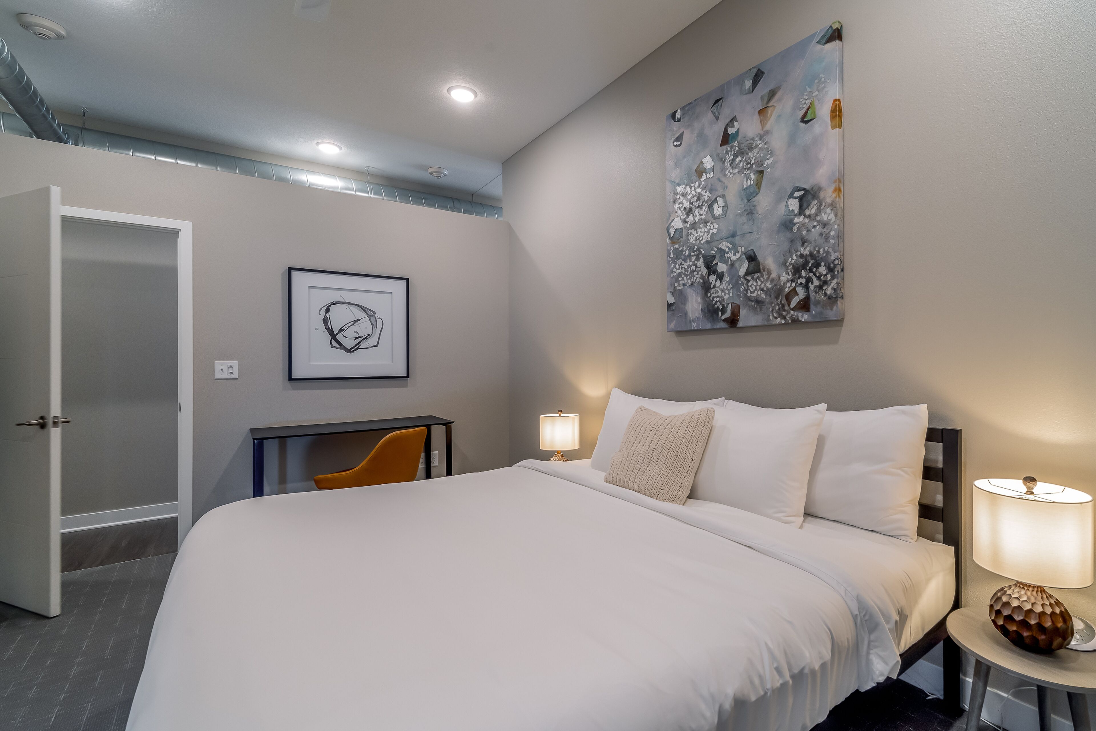 Een hotelkamer met een groot bed, twee bedlampen, een bureau met een stoel, een schilderij aan de muur en een deur die leidt naar een andere kamer.