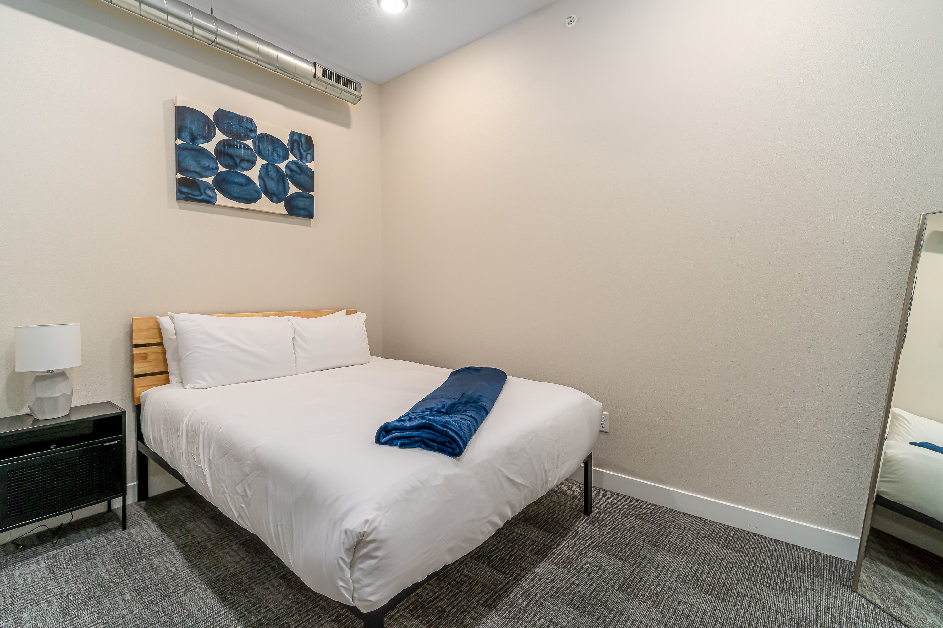 A hotel room with a neatly made bed, a bedside table with a lamp, a wall-mounted air conditioner, and a painting above the bed.