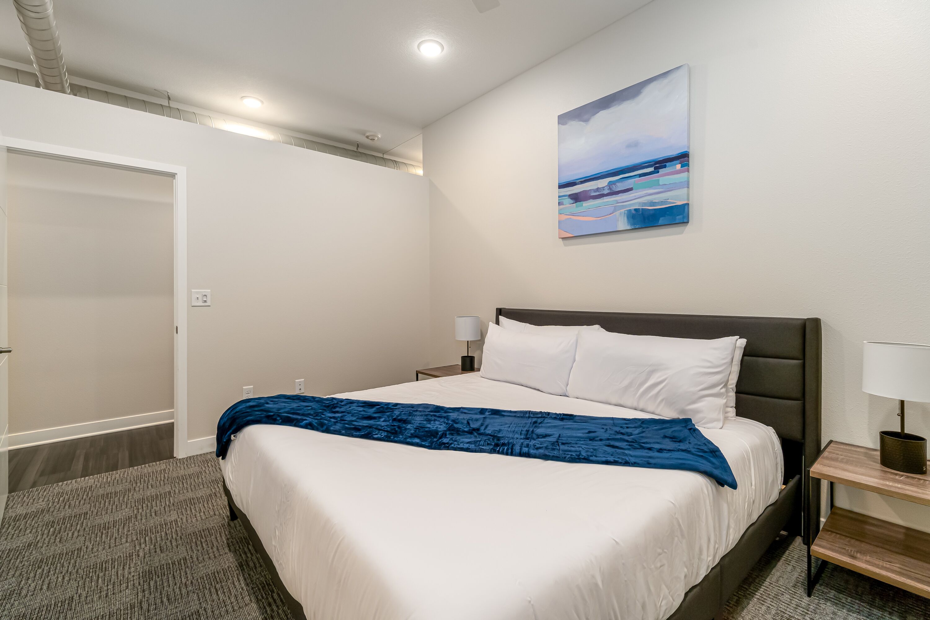 A neatly made bed with white linens and a blue throw blanket, a bedside table with a lamp, and a painting of a landscape on the wall.