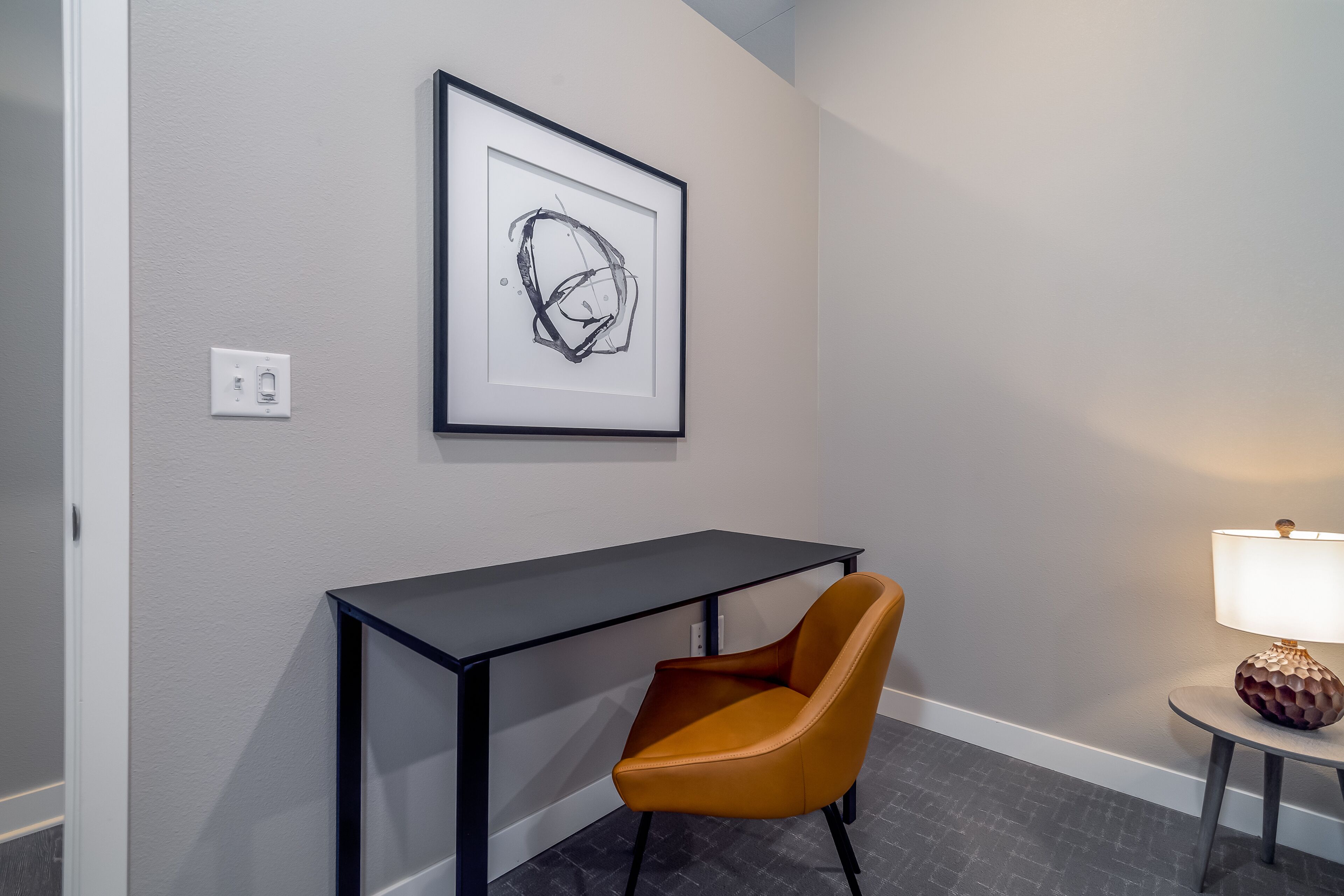 A room with a desk, a framed abstract artwork, a chair, and a lamp.