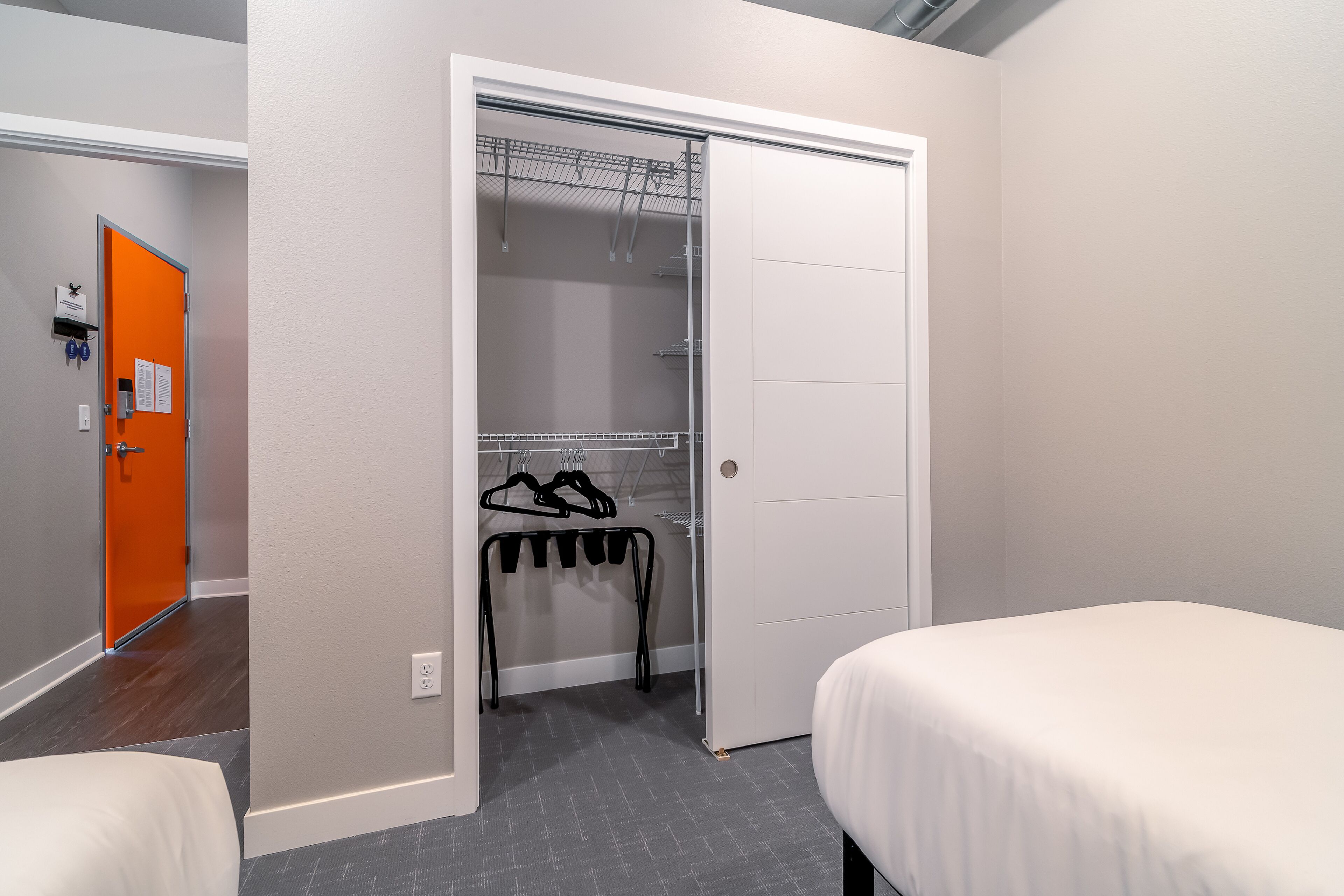 A hotel room with a grey carpet, a white bed, a closet with a metal rack, and an orange door.