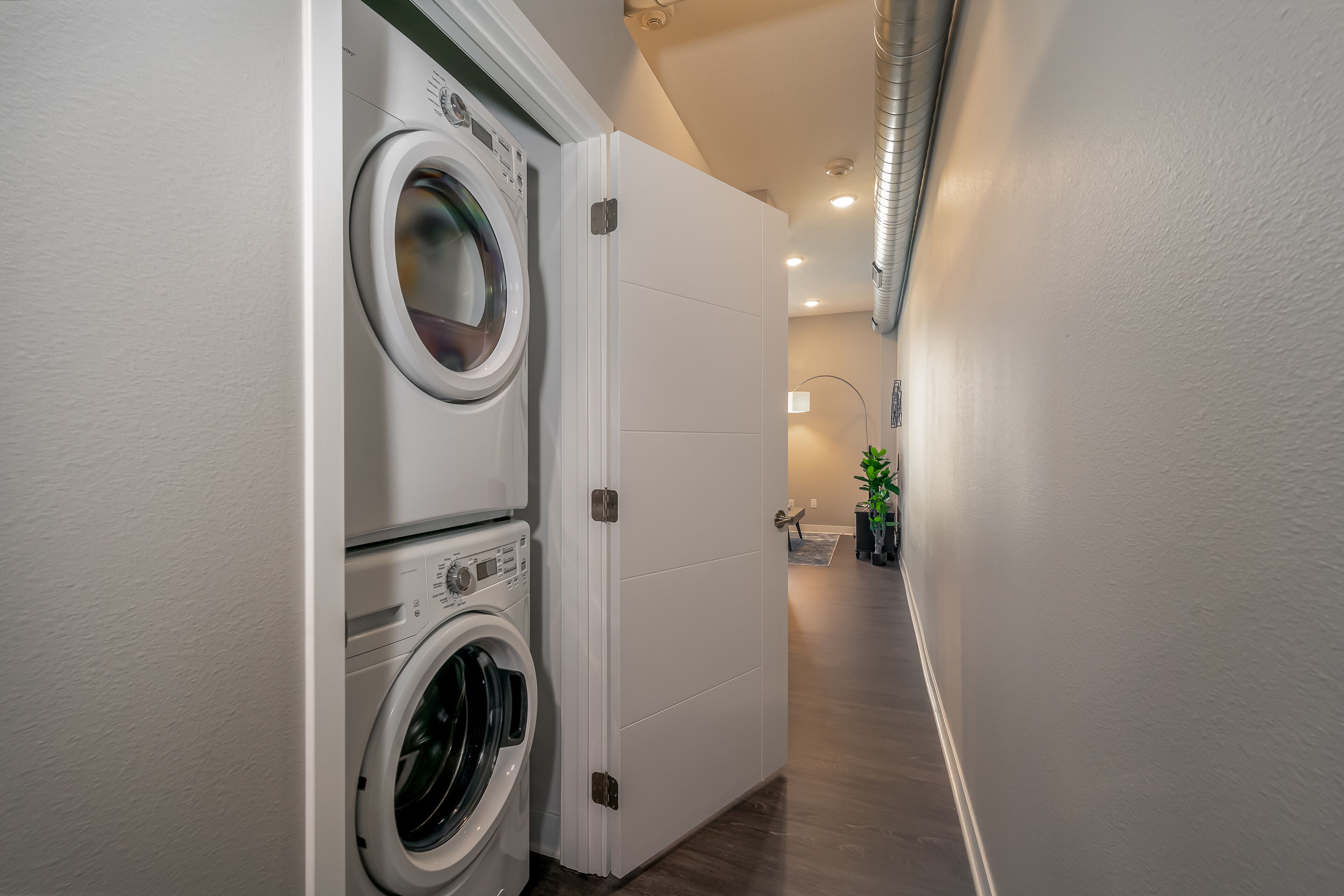 A laundry room with a front-loading washer and dryer, a white door, and a hallway leading to a bathroom with a sink and a plant.