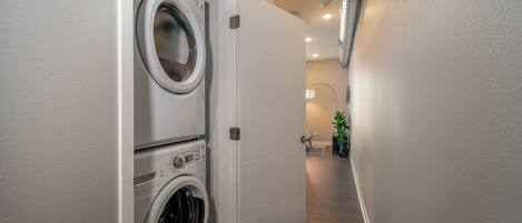 A laundry room with a front-loading washer and dryer, a white door, and a hallway leading to a bathroom with a sink and a plant.