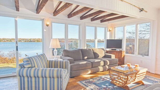 A spacious living room with a large window overlooking a lake, a sofa, a striped armchair, and a coffee table. - Waterfront amazing views near beaches! (Eastham)