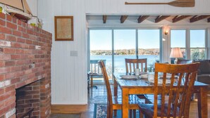 A dining area with a brick fireplace, wooden dining table, and chairs, with a view of the water outside. - Waterfront amazing views near beaches! (Eastham)