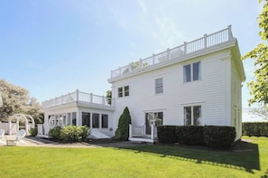 A two-story white building with a balcony on the upper floor, a covered porch on the ground floor, and a well-maintained lawn. - Quintessential Captain's House w/ Water Views! (Eastham)