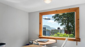 Una habitación con una mesa y sillas de madera, un ventanal con vistas a casas y follaje, y una pared blanca.