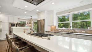 Una cocina amplia con una isla grande, armarios blancos, electrodomésticos de acero inoxidable y vistas a árboles a través de amplias ventanas.
