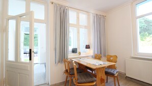 Un comedor con mesa y sillas de madera, ventanas amplias con cortinas y vista a follaje exterior.