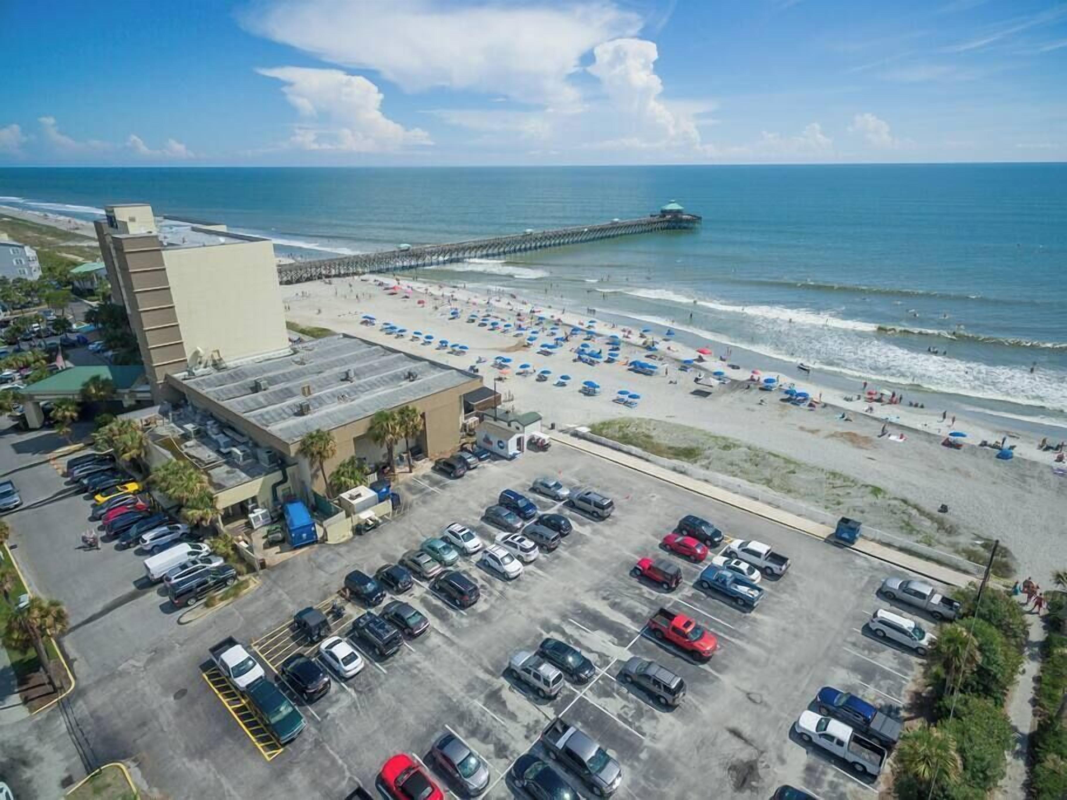 An aerial view of a beachside parking lot with a pier extending into the ocean, adjacent to a beach with umbrellas and people.