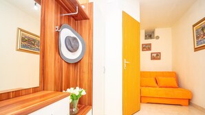 A room with a wooden vanity, a white dresser, a mirror, a hat, and a vase with flowers. There is an orange sofa and a door leading to another room.