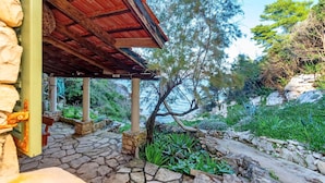 A stone-paved patio with a wooden pergola, surrounded by lush greenery and a view of the sea.