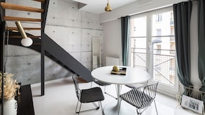 A modern living space with a round dining table, black wireframe chairs, a white vase with dried flowers, a wooden staircase, and a large window with curtains. - Jad'Or - Jolie Duplex avec parking privé (Nantes)