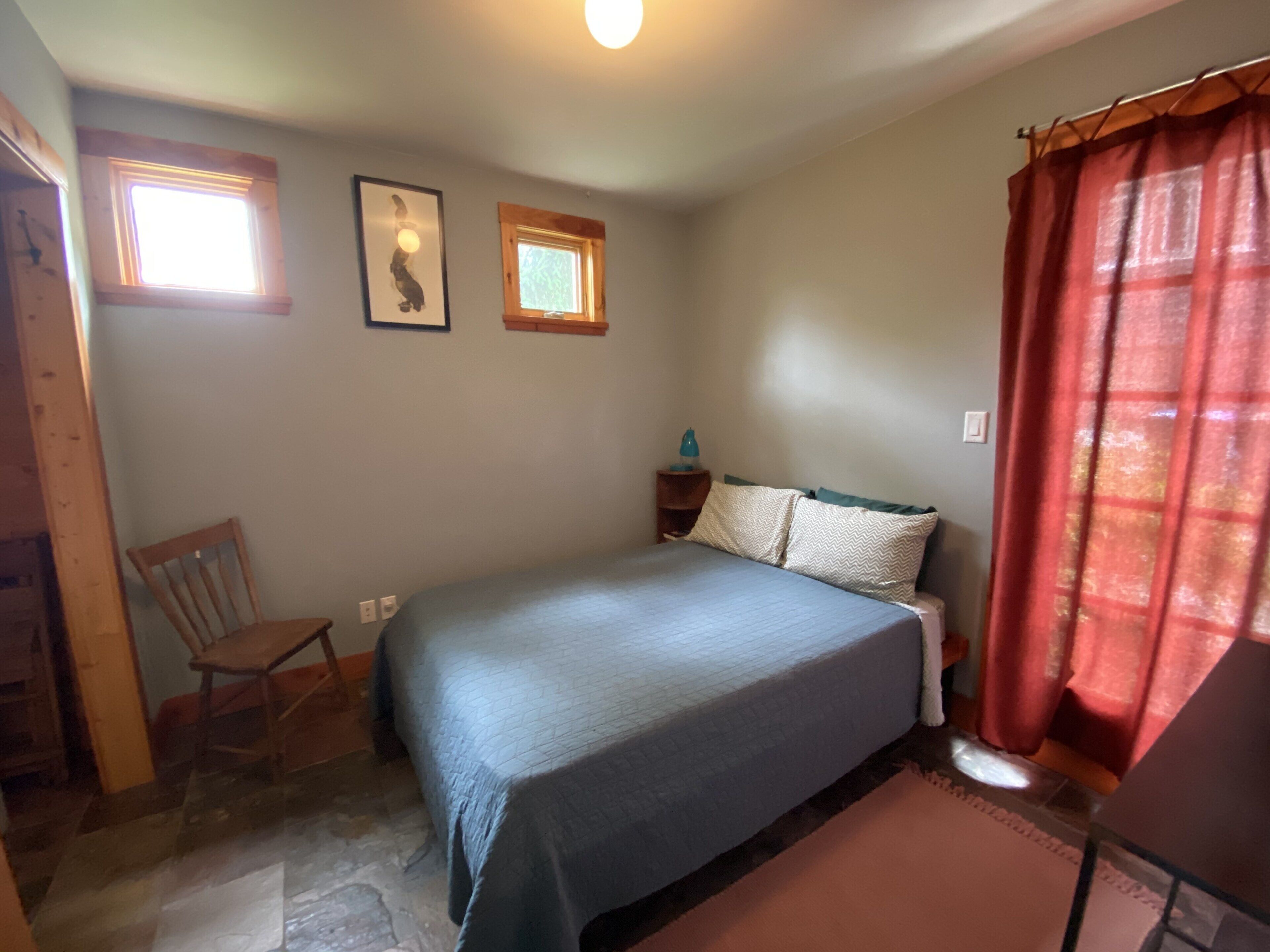 Ein Schlafzimmer mit einem Bett, einem Sessel, einem Nachttisch und zwei Fenstern.