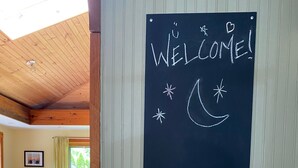 A room with a wooden ceiling, a chalkboard with "WELCOME!" and a crescent moon, a table with a bottle, and a view of a dining area. - Charming & cozy in-town retreat in the heart of New Buffalo (New Buffalo)