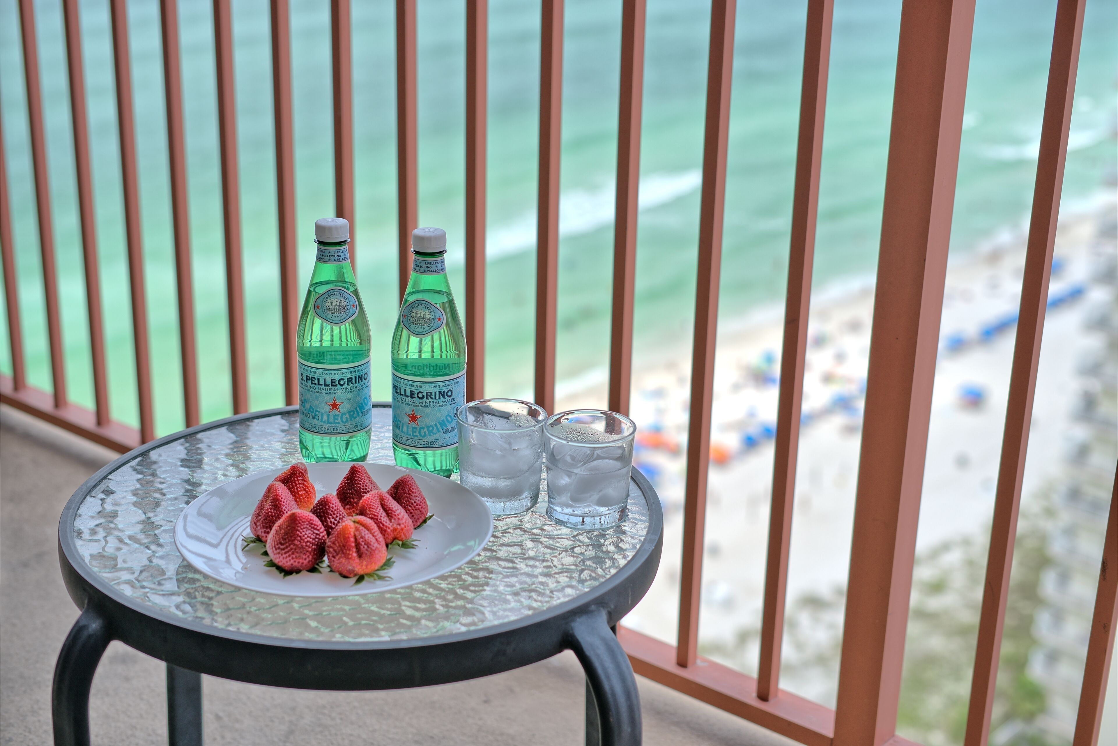 A table with two bottles of Pellegrino water, two glasses of water, and a plate with strawberries on a balcony overlooking the beach.