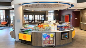 A self-service beverage station with a variety of drinks and snacks, a seating area with a chair, and a counter with a digital display. - Tru By Hilton Baltimore Harbor East, MD (Baltimore)