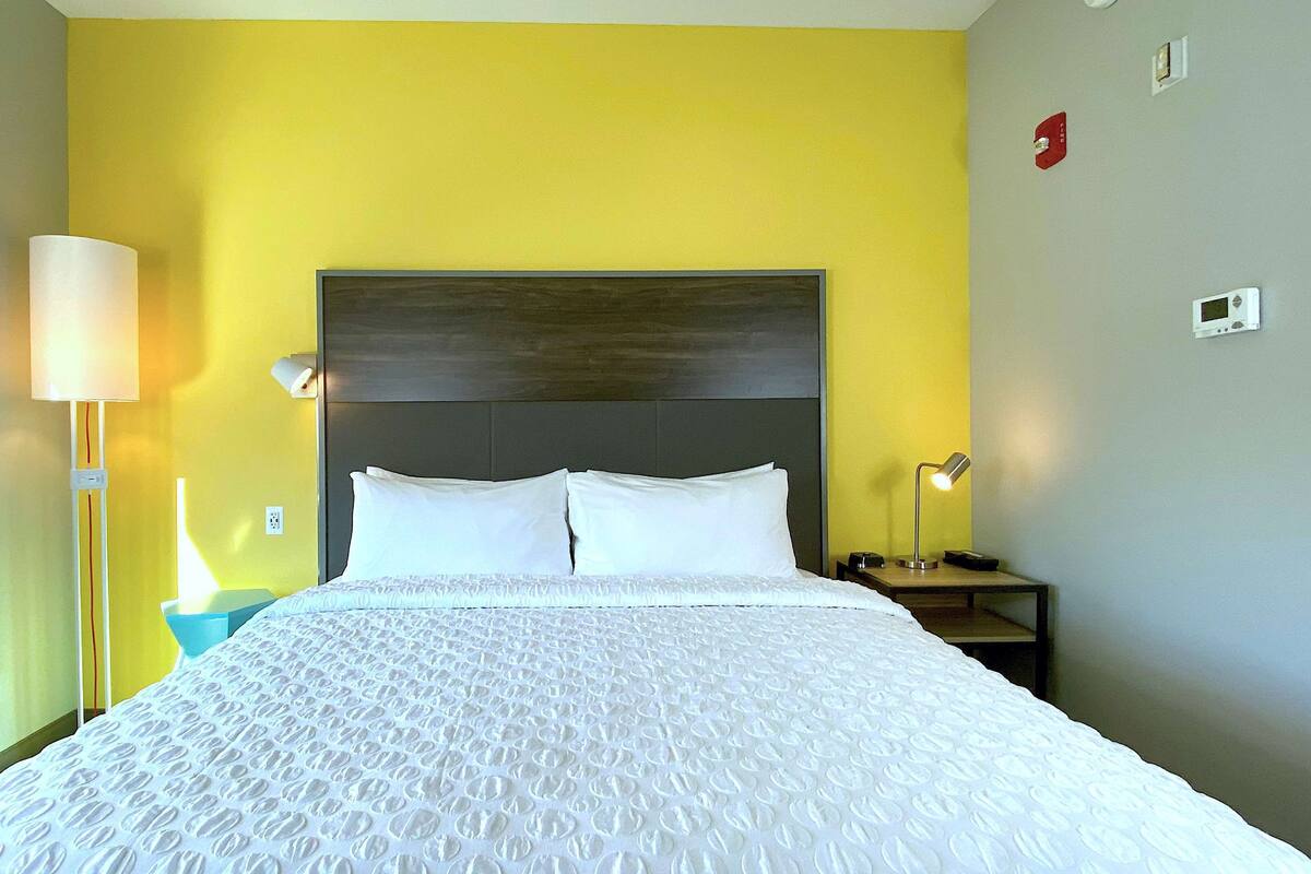 Ein Hotelzimmer mit Bett, Nachttisch, Nachttischlampe und Wandthermostat.