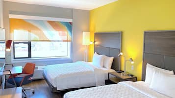 A hotel room with two beds, a desk, a chair, and a window with a view of buildings.