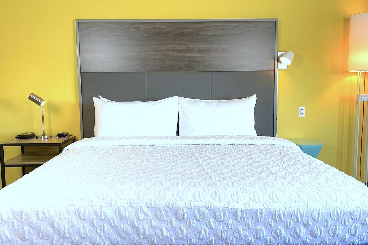 Ein Hotelzimmer mit einem großen Bett, einem Nachttisch mit Lampe sowie einem Beistelltisch mit Telefon und Ladegerät.