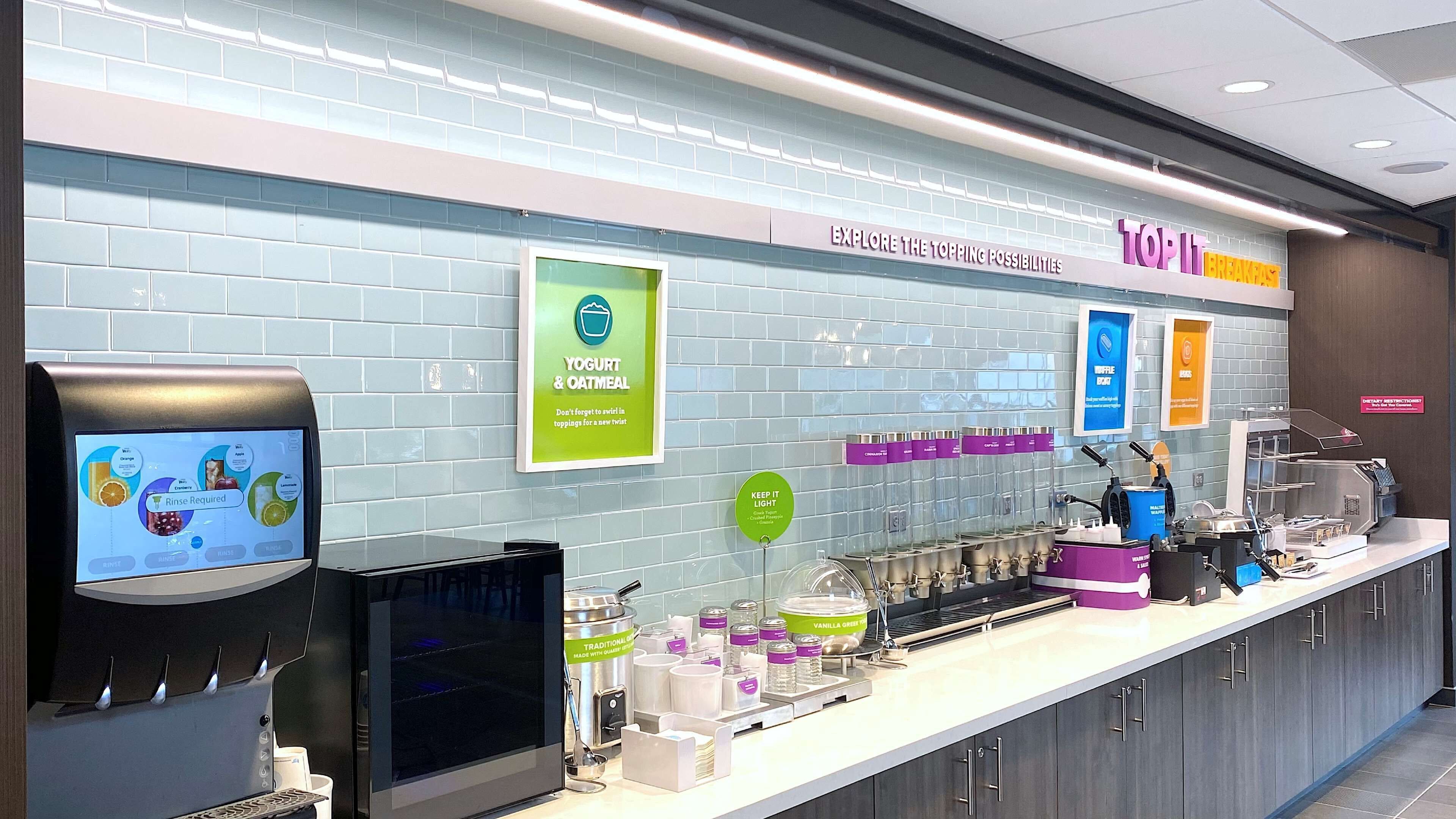 Un área de desayuno autoservicio con menú digital, opciones de yogur y avena, estación de gofres y una máquina de café.