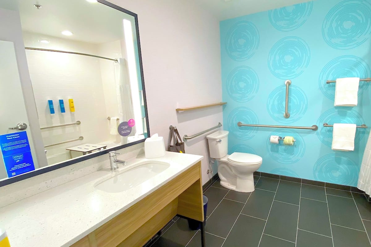 Ein Badezimmer mit weißem Waschbecken, Spiegel und Toilette. Es gibt Handläufe und einen Handtuchhalter.