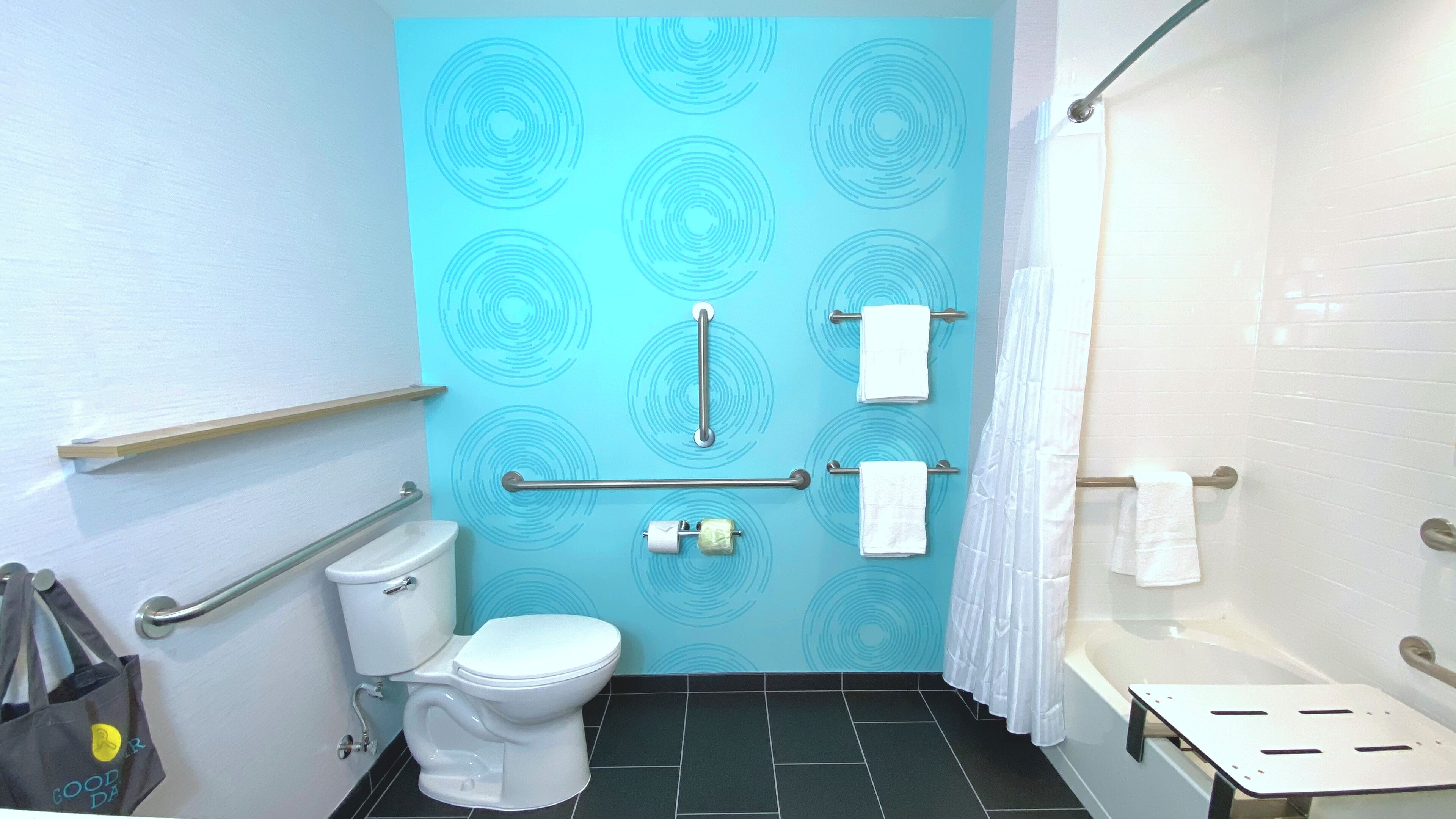 Un baño con pared azul con estampado, inodoro blanco, barra de apoyo, toallero, cortina de ducha y un pequeño banco.