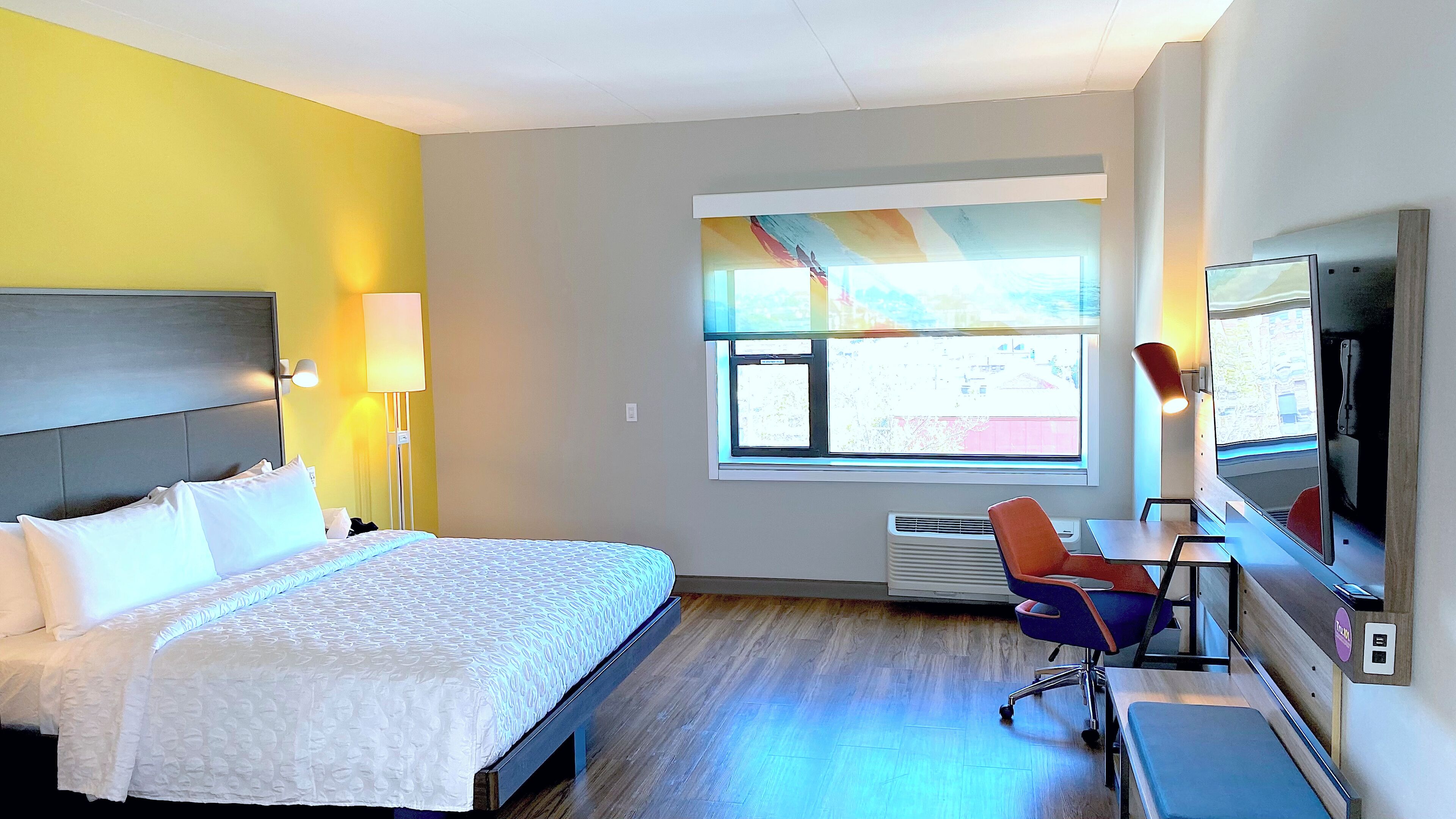 A hotel room with a bed, a desk with a chair, a large window, and a TV.