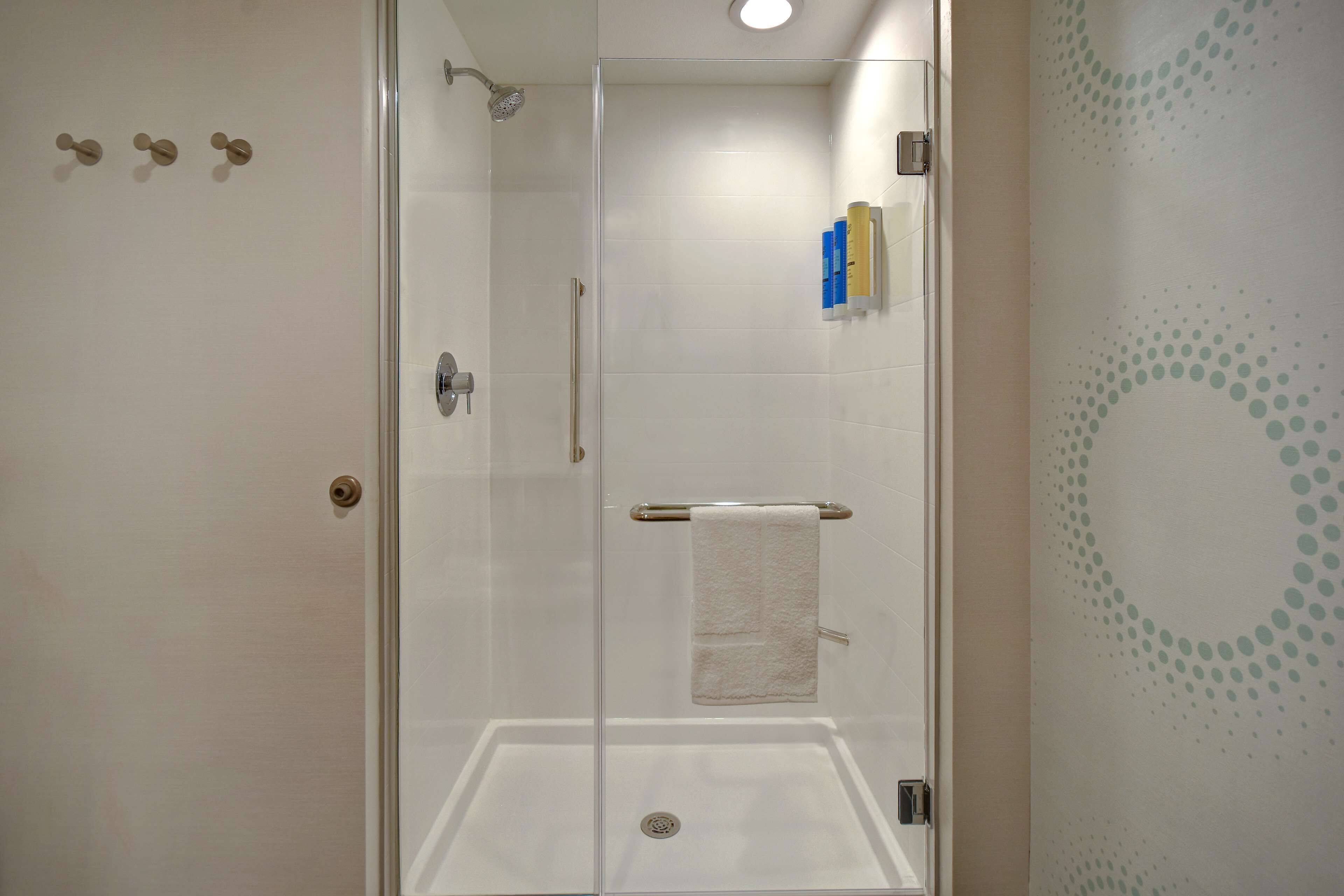Ducha con cerramiento de vidrio, una toalla blanca colgada en un perchero, una puerta con pomo redondo y una pared con diseño circular.