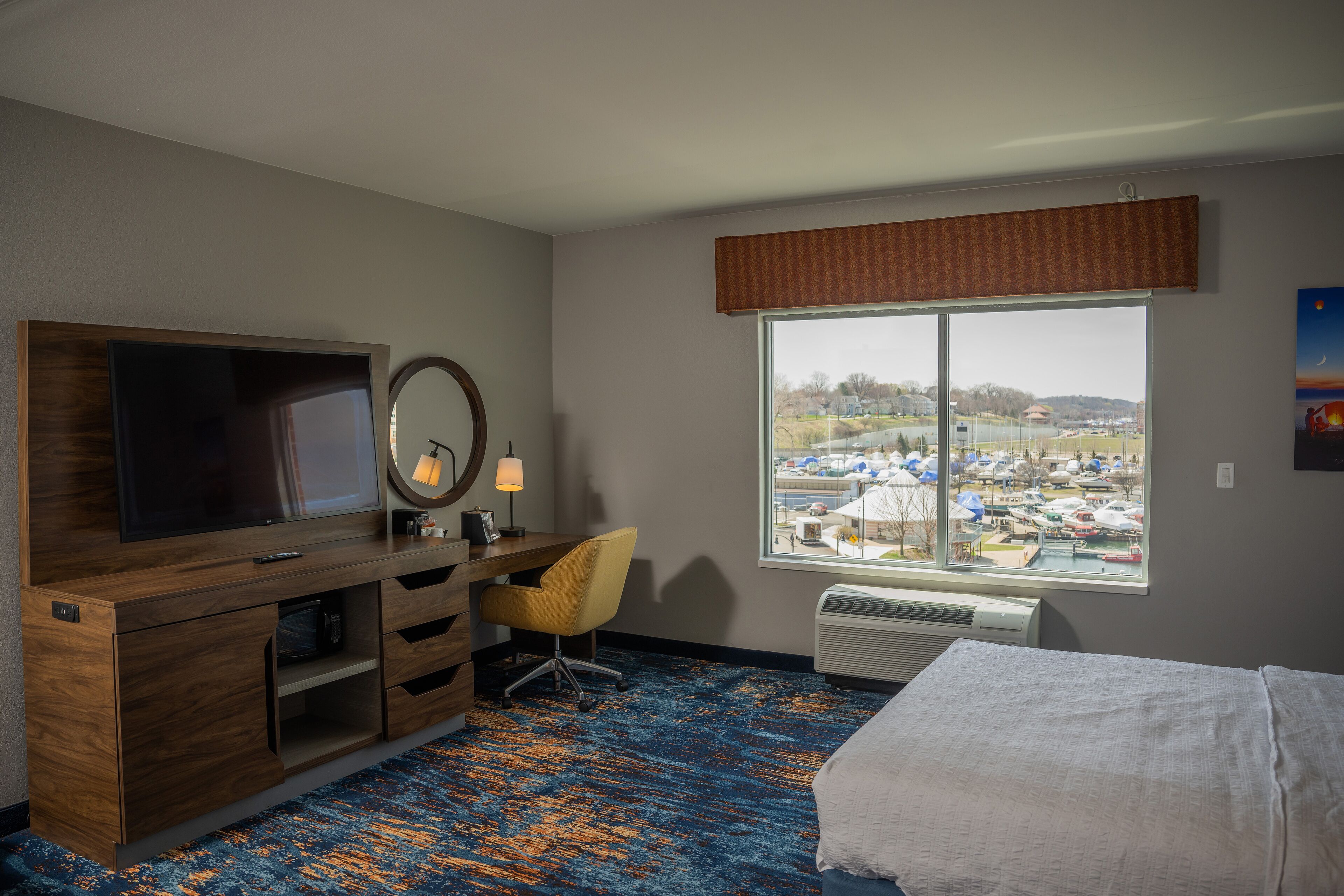A hotel room with a large window, a bed, a desk with a chair, a TV, and a mirror.