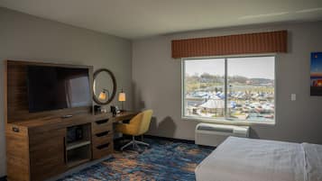 A hotel room with a large window, a bed, a desk with a chair, a TV, and a mirror.