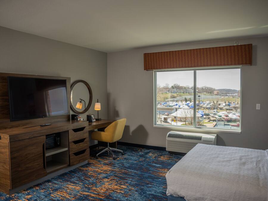 A hotel room with a large window, a bed, a desk with a chair, a TV, and a mirror.