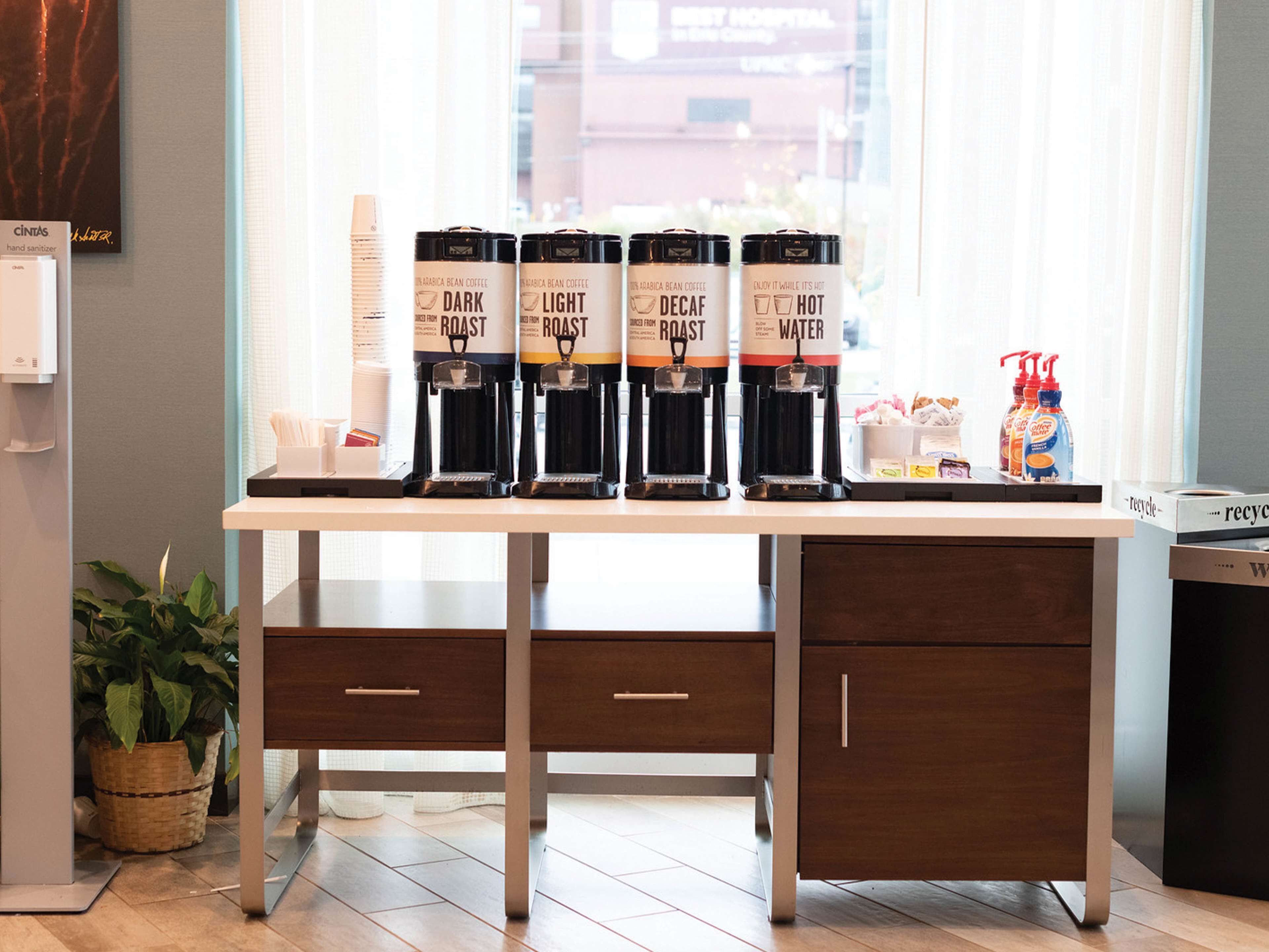 Un espace dédié au café est aménagé avec quatre distributeurs de café étiquetés « FORTE », « DOUCE », « DÉCAFÉINÉ » et « CHAUD ». On y trouve également des bouteilles de gel hydroalcoolique et une poubelle de recyclage sur la table.