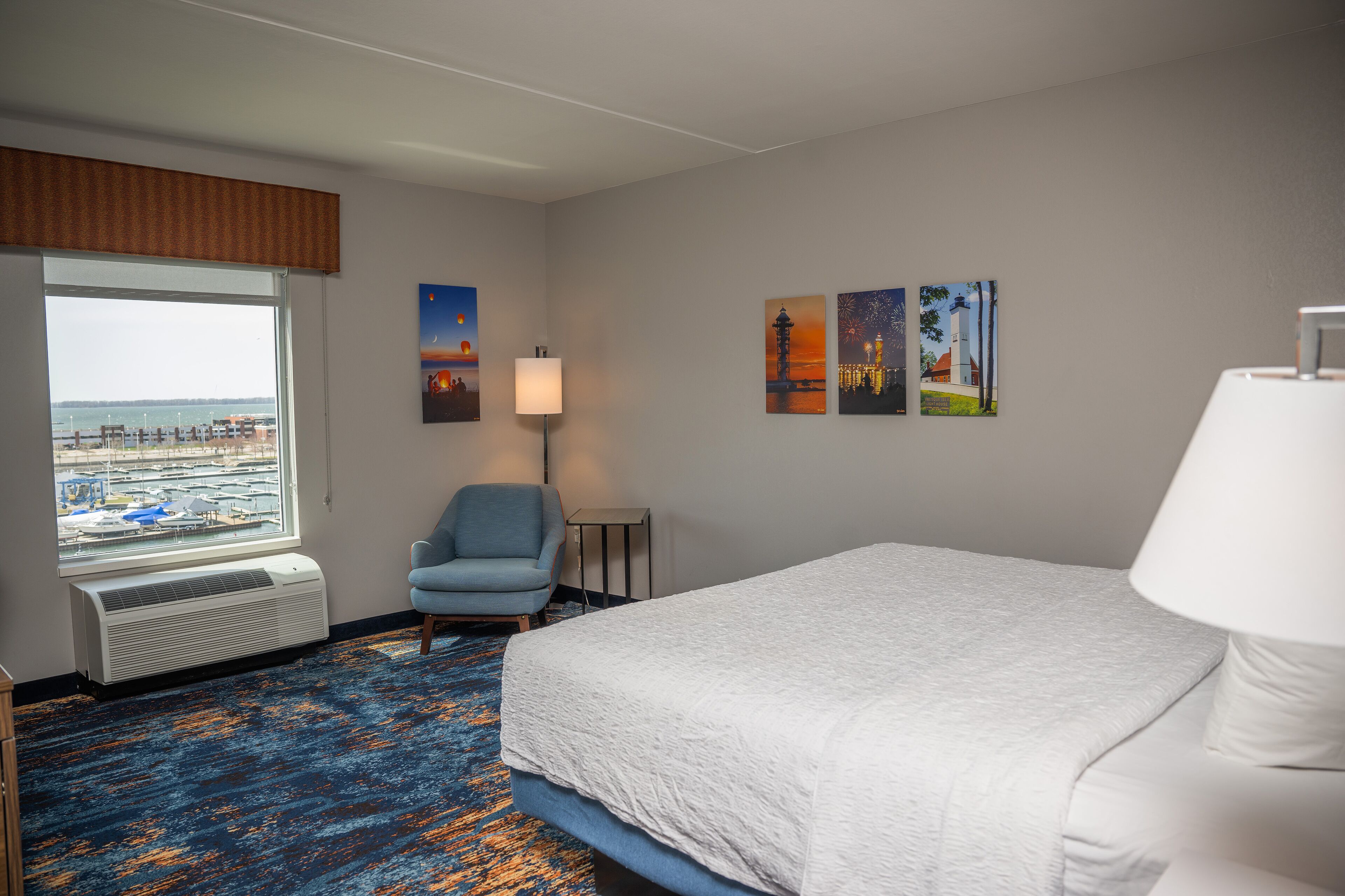 A hotel room with a large bed, a chair, a desk, and a window with a view of the beach and cityscape.